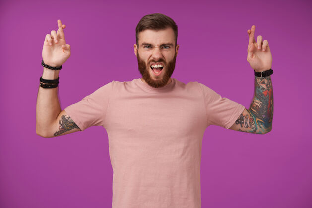 模特年轻的纹身黑发男子的肖像 胡子举起交叉的手指在好运的姿态 皱着眉头 保持张嘴 而紫色的姿势穿着室内身体