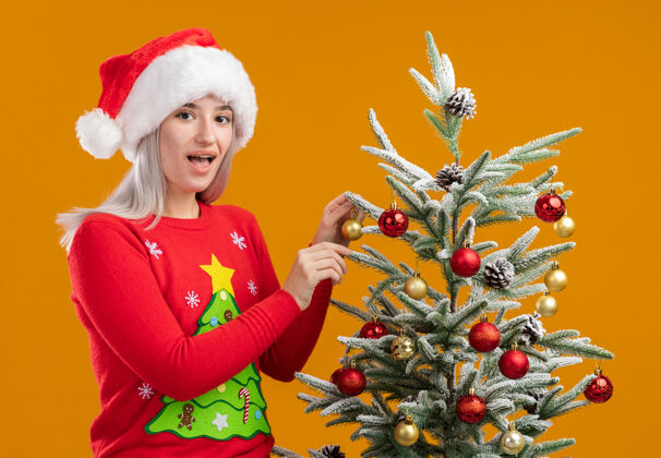 积极年轻的金发女郎穿着圣诞毛衣 戴着圣诞帽装饰着圣诞树快乐而积极的微笑着快乐的圣诞树圣诞帽子金发