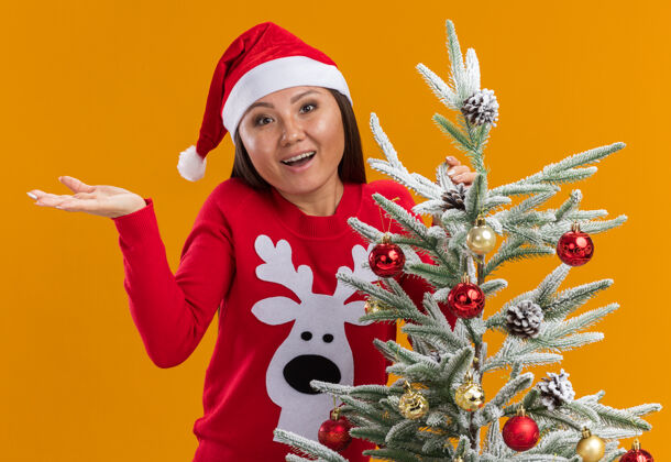 亚洲人惊讶的年轻亚洲女孩戴着圣诞帽和毛衣站在圣诞树旁摊开手孤立在橙色背景女孩圣诞附近
