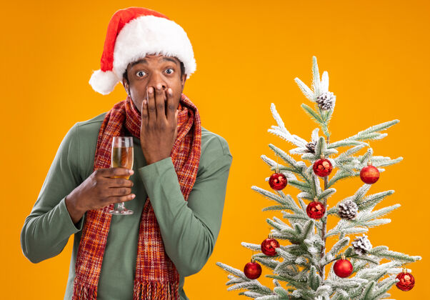 手戴着圣诞帽 围着围巾的非洲裔美国人举着一杯香槟看着摄像机震惊地用手捂住嘴巴 旁边是橙色背景下的圣诞树站着树男人