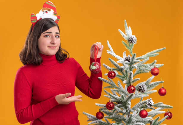 毛衣困惑的小女孩穿着圣诞毛衣戴着有趣的头带旁边的圣诞树橙色背景看头带圣诞节