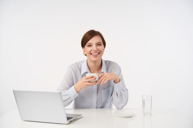 女性快乐的棕色头发的年轻女子 随意的发型 在工作日一边喝咖啡一边开心地笑着 穿着蓝色衬衫一边摆着白色的姿势自然正式员工