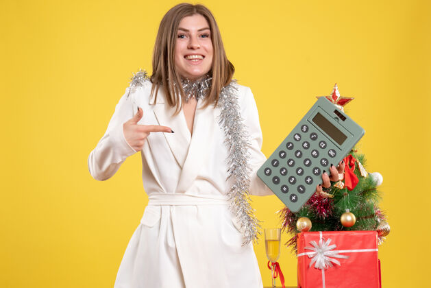 站着正面图：女医生站在黄色桌子上 手里拿着计算器 桌子上放着圣诞树和礼品盒快乐女医生桌子