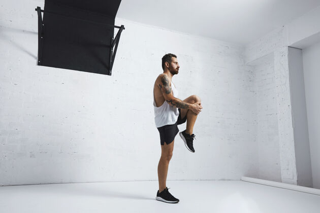 工作运动型年轻人做抬腿动作 伸直双腿 看向右侧 隔离在白砖墙旁的拉杆上健康身体体操