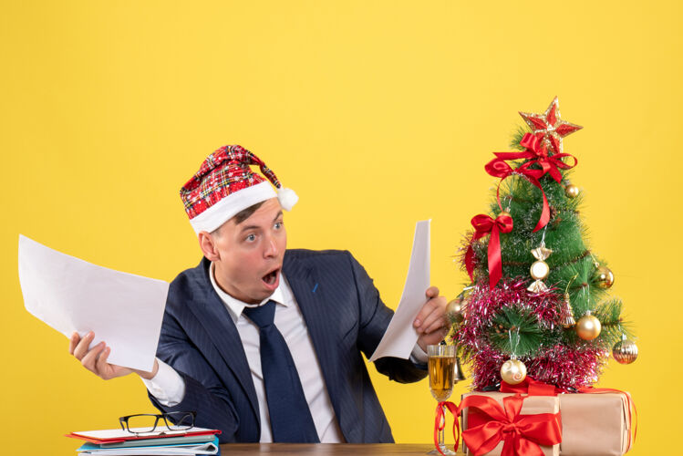 坐着大眼睛男人坐在圣诞树旁的桌子旁检查文件和黄色礼物的前视图商人支票商人