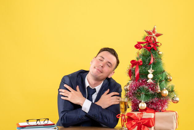新郎睡意朦胧的男人交叉双手坐在圣诞树旁的桌子前 黄色的礼物坐着桌子瞌睡