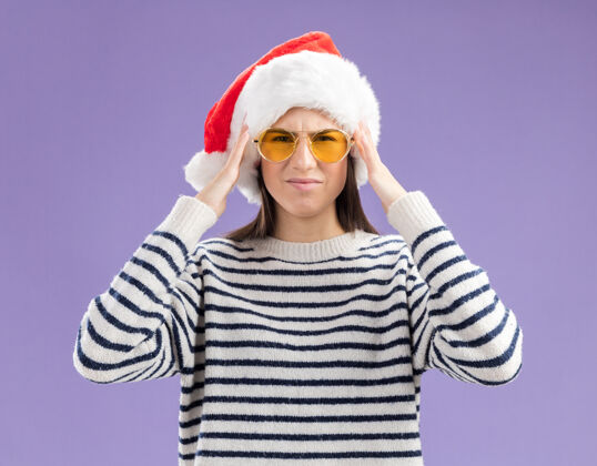 眼镜戴着太阳眼镜 戴着圣诞帽 心疼的白种年轻女孩把双手放在头上 紫色背景上 留有复制空间头圣诞节放