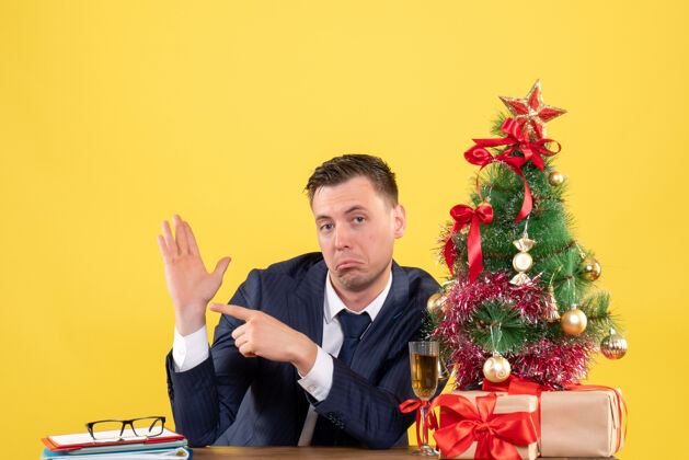 黄色好奇的男人手指着他的手坐在圣诞树和黄色礼物旁边的桌子的前视图桌子好奇的人树