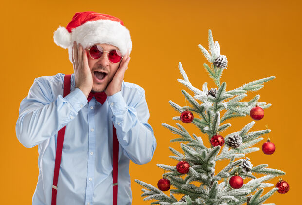 蝴蝶结站在橙色墙上的圣诞树旁 戴着圣诞帽 戴着红眼镜 系着吊带领结的年轻人惊讶不已圣诞节帽子近
