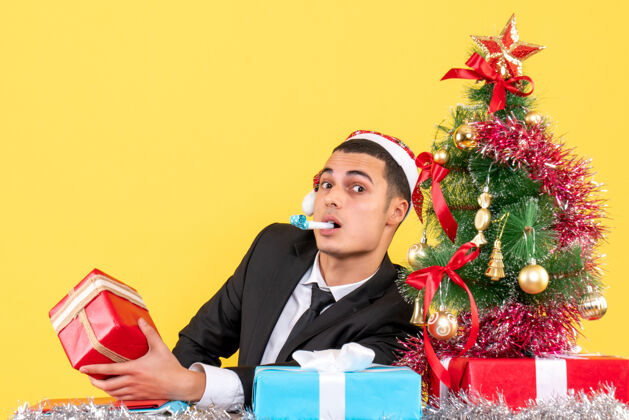 男人前视图惊讶的男子戴着圣诞帽坐在桌子旁拿着礼物用噪音制造圣诞树和礼物礼物商人惊讶