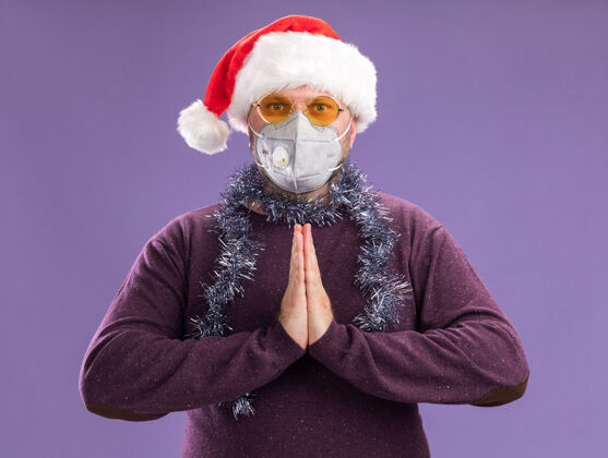 脖子中年男子戴着圣诞老人帽 脖子上戴着金属丝花环 戴着眼镜和防护面具 手在紫色的墙上保持着隔离保管花环圣诞老人