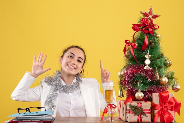 人前视图：女医生坐在黄色背景的桌子后面 带着圣诞树和礼品盒插花健康圣诞节