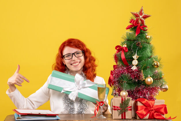 礼物前视图：女医生坐在黄色背景上 手里拿着圣诞礼物和圣诞树坐着背景女医生