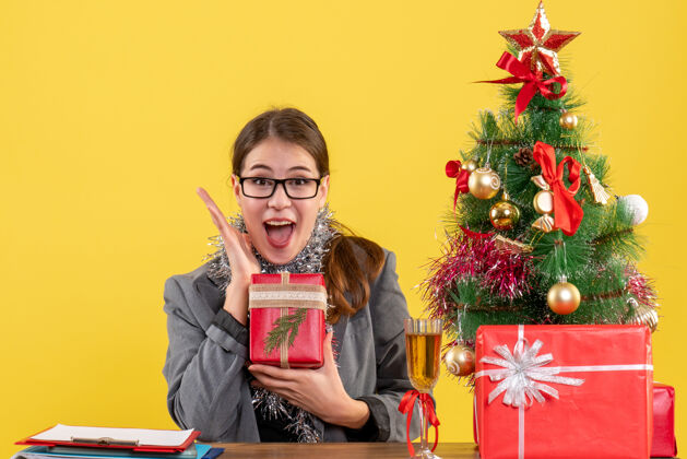 鸡尾酒前视图欣喜若狂的女孩戴着眼镜坐在桌旁 展示她的幸福圣诞树和鸡尾酒礼物展示坐着幸福