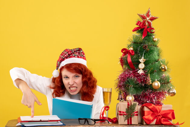 医生正面图：女医生坐在桌子后面 手里拿着文件 背景是黄色的圣诞树和礼盒前面情感圣诞节