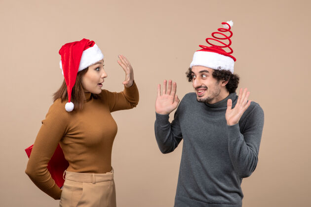 圣诞节新年气氛喜庆的概念与有趣可爱的情侣戴着红色圣诞老人帽女孩隐藏礼物背后的灰色圣诞老人帽子帽子