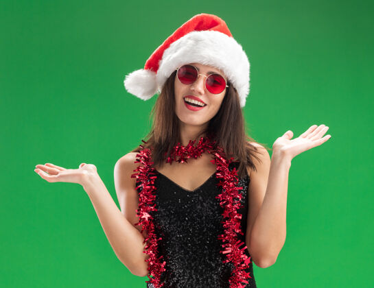 脖子微笑着的年轻漂亮女孩戴着圣诞帽戴着眼镜脖子上戴着花环摊开双手隔离在绿色的墙上蔓延穿着圣诞节