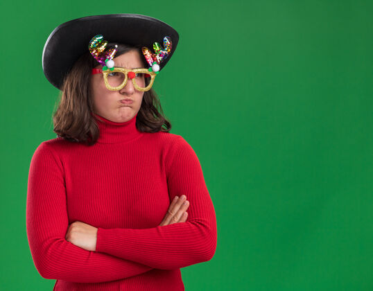 不高兴穿着红毛衣的年轻女孩戴着滑稽的眼镜和黑帽子 不高兴地向一边望去 双臂交叉着 脸颊朝天 站在绿色的墙上帽子穿交叉