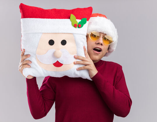 年轻惊讶的年轻人戴着圣诞帽戴着眼镜抱着圣诞枕头孤立在白色的墙上枕头圣诞节帽子