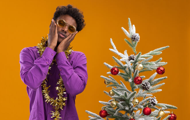 保持困惑的美国黑人年轻人戴着眼镜 脖子上戴着金箔花环 站在装饰过的圣诞树旁 手放在脸上 孤立地站在橙色的墙上靠近脖子圣诞节