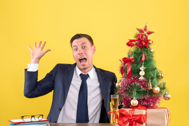 男正面图是坐在桌子旁的大眼睛男人在圣诞树旁打招呼 黄色的礼物西装会议坐着