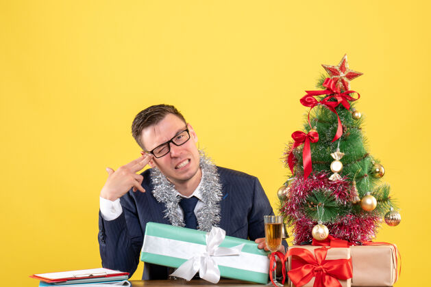 圣诞节前视图的快乐男子把手指枪到他的太阳穴坐在桌旁圣诞树和黄色的礼物礼物快乐男人树