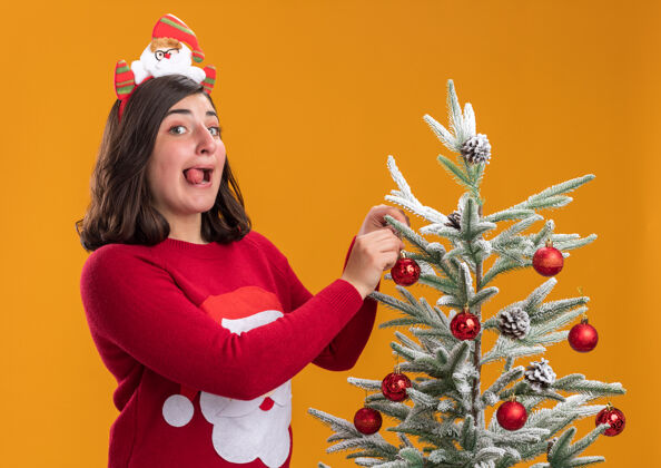 圣诞节穿着圣诞毛衣 戴着滑稽的头带 快乐快乐的小女孩站在圣诞树旁 圣诞球挂在橙色的墙上穿站毛衣