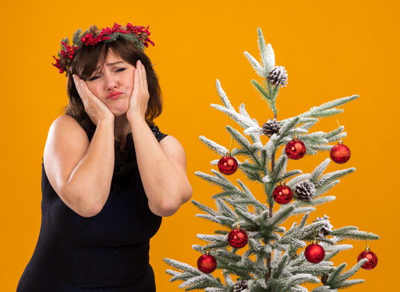 围着悲伤的中年妇女戴着圣诞花环 脖子上戴着金属丝花环 站在装饰好的圣诞树旁 手放在脸上 隔离在橙色的墙上靠近保持戴着