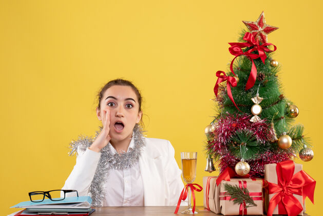 健康前视图：女医生坐在黄色背景的桌子后面 带着圣诞树和礼品盒导演肖像成人