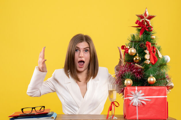 微笑正面图：女医生坐在黄色背景的桌子前 放着圣诞树和礼品盒人办公室背景