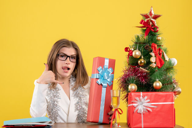 女医生正面图：女医生坐在桌子前 拿着礼物 黄色桌子上的圣诞树和礼盒树前面人