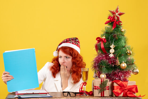 背景正面图：女医生坐在桌子后面 拿着黄色背景的文件 还有圣诞树和礼品盒肖像抱着餐桌