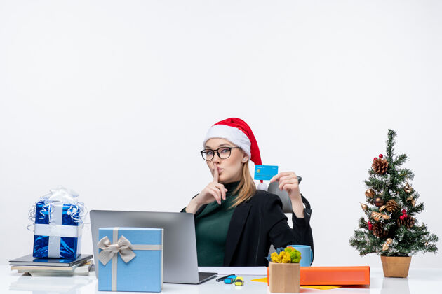 桌子戴着圣诞老人帽 戴着眼镜的女商人坐在桌子旁 手里拿着圣诞礼物和银行卡 在办公室里做着沉默的手势电脑手势佩戴