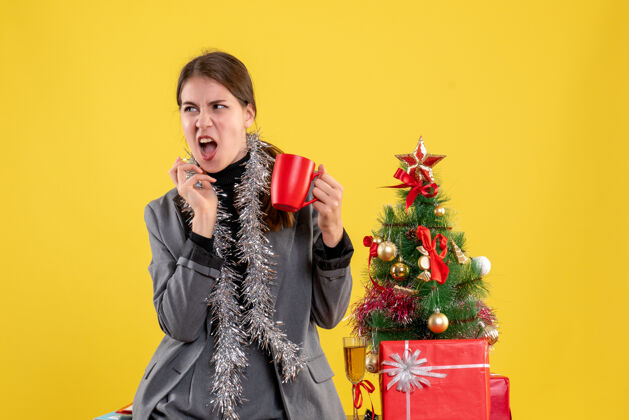 成人正面图：年轻女孩张开嘴 拿着圣诞树旁的红色杯子和鸡尾酒嘴肖像桌子