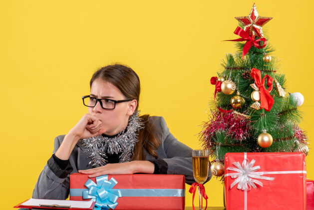 坐着前视图带着眼镜的体贴女孩坐在桌子旁思考圣诞树和鸡尾酒礼物桌子微笑生活