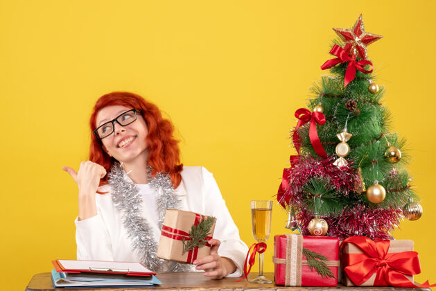 圣诞节前视图：女医生坐在桌子后面 黄色背景上有圣诞礼物快乐护士礼物