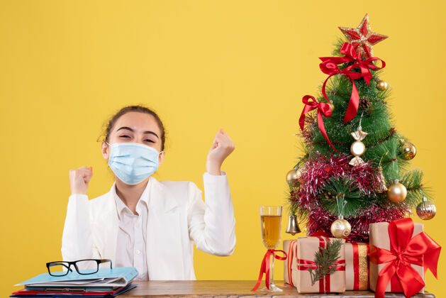 人正面图女医生戴着无菌口罩坐在黄色背景上 带着圣诞树和礼品盒专业男性外套