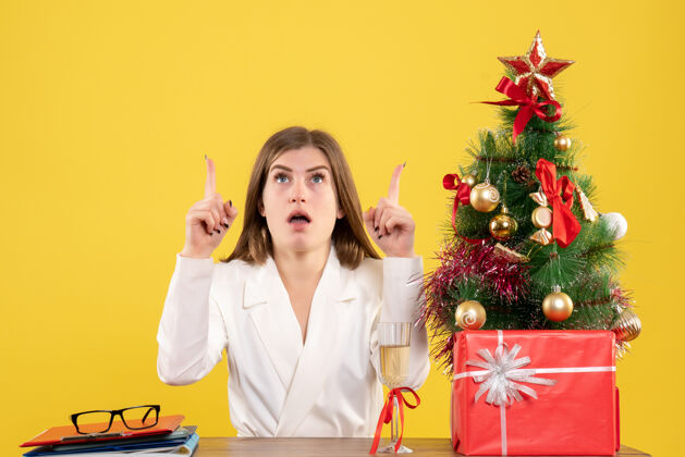 职业正面图：女医生坐在黄色背景的桌子前 放着圣诞树和礼品盒医院女性人