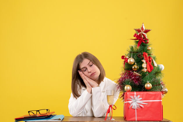 人正面图：女医生坐在黄色背景的桌子前 放着圣诞树和礼品盒坐着医生家