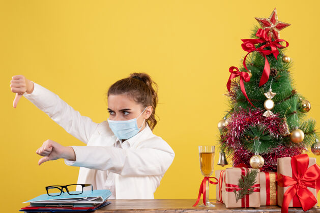 无菌正面图女医生戴着黄色背景的无菌口罩坐在圣诞树和礼盒里工作坐着男性