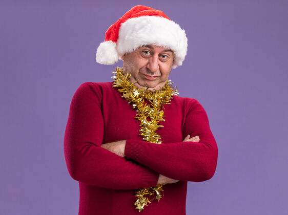 圣诞节中年男子戴着圣诞老人帽子 脖子上戴着金属丝 双臂交叉站在紫色背景上 带着怀疑的表情看着相机年龄交叉站着