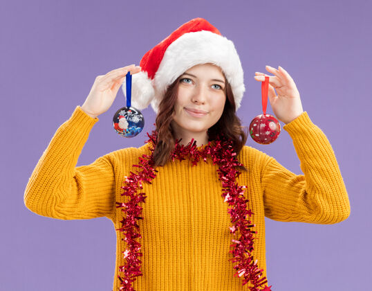 球年轻的斯拉夫女孩戴着圣诞帽 脖子上戴着花环 手里拿着玻璃球饰品 背景是紫色的 有复制空间新年斯拉夫装饰品