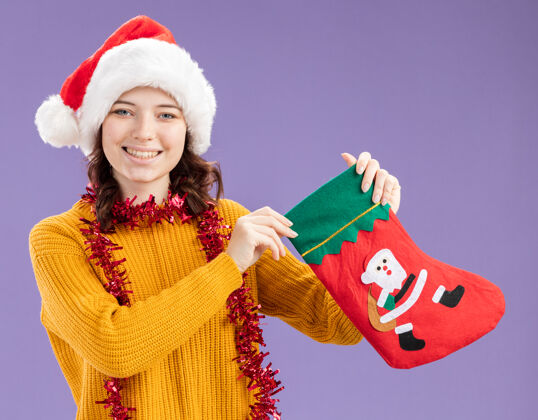圣诞快乐微笑着的年轻斯拉夫女孩 戴着圣诞帽 脖子上戴着花环 手里拿着圣诞长袜 背景是紫色 有复制空间围着圣诞节微笑