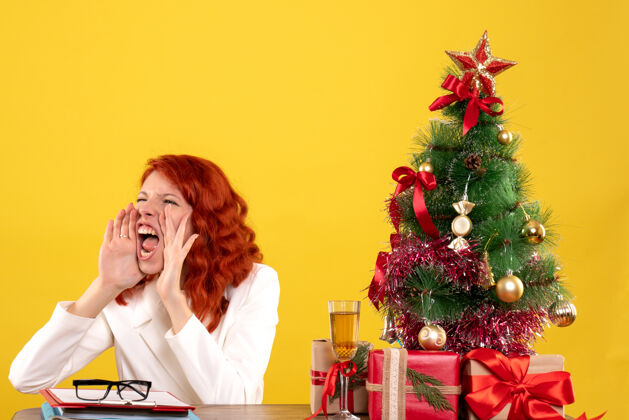 女性正面图：女医生坐在桌子后面 手里拿着圣诞礼物 黄色背景上的圣诞树在呼唤前面快乐医生