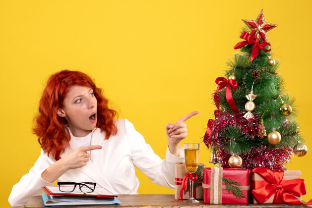 微笑正面图：女医生坐在桌子后面 拿着圣诞礼物 黄色背景上的树成人女性前面