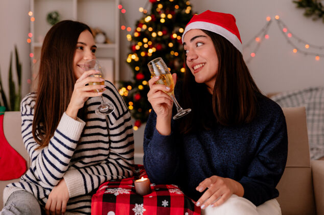 快乐高兴的年轻女孩们拿着香槟酒杯 坐在扶手椅上互相对视 在家里享受圣诞节时光家漂亮圣诞节