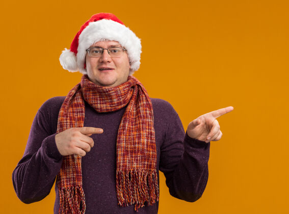 高兴一个戴着眼镜 戴着圣诞帽 脖子上围着围巾 看着摄像机指着橙色背景上孤立的一边的男人眼镜圣诞老人橙色