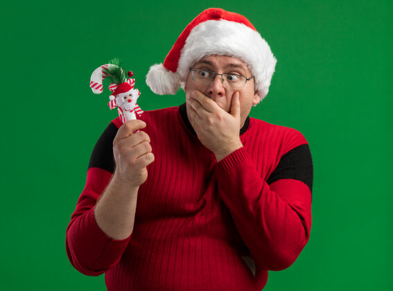 关心戴着眼镜 戴着圣诞老人帽子 手放在嘴上 手拿着糖果 手捧着藤条 看着绿色的背景眼镜快乐抱