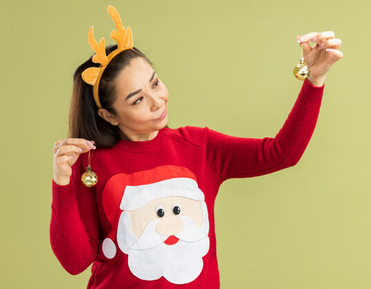 年轻穿着红色圣诞毛衣的年轻女子 戴着滑稽的带鹿角的边 手里拿着圣诞球 看上去困惑不解 心中充满疑惑角圣诞快乐困惑