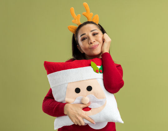 新年穿着红色圣诞毛衣的快乐年轻女子 戴着滑稽的鹿角边 抱着圣诞枕头 面带微笑拿着微笑枕头